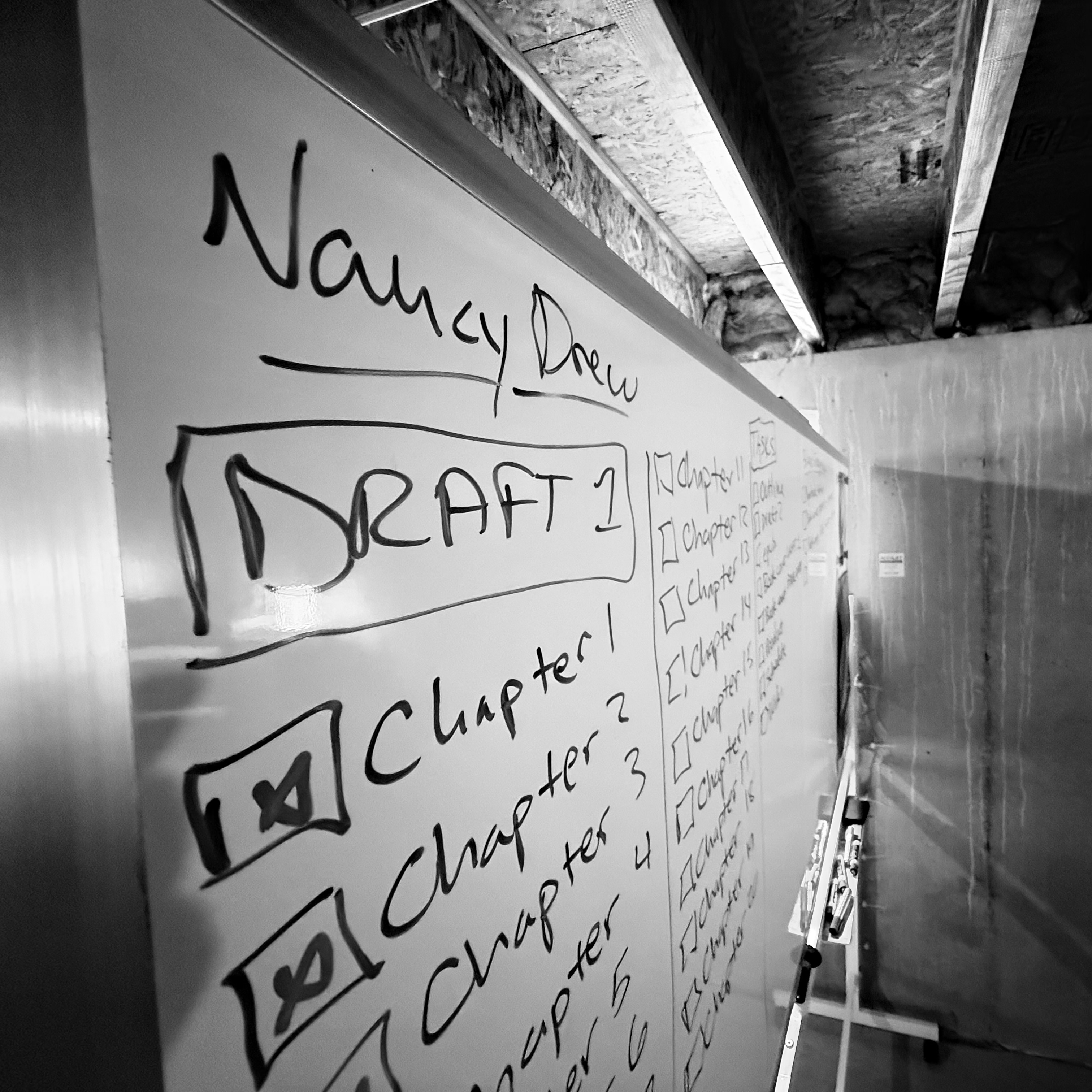 A black and white image of a long white board that is on wheels in a basement. There is a task list written on the board under the header that reads "Nancy Drew". The tasks are various chapters in the first draft of the work. The first three chapters are checked off as completed with an X.